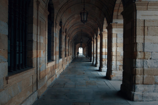 A long stone arcade corridor with repeating arches and columns, dark barred windows on the left, and a bright opening at the far end.