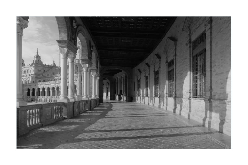 Black and white architectural photograph of a long covered arcade corridor with columns, tiled floor, and a distant domed building, presented as a square print with a thin white border.