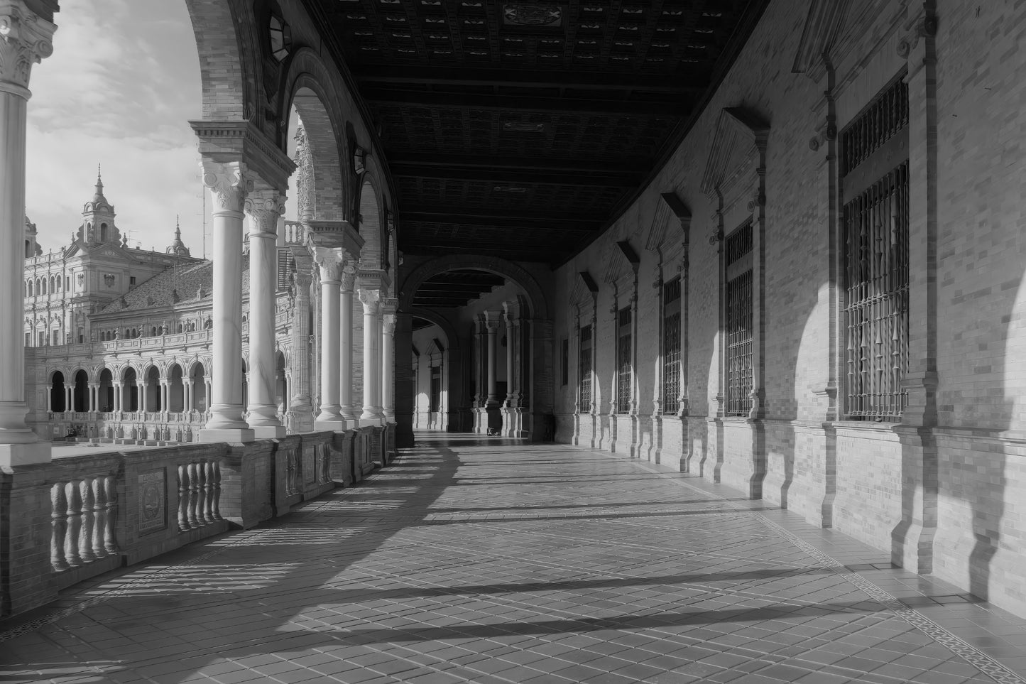Black and white photograph of a grand architectural colonnade with arches, columns, tiled floor, and a distant ornate building.