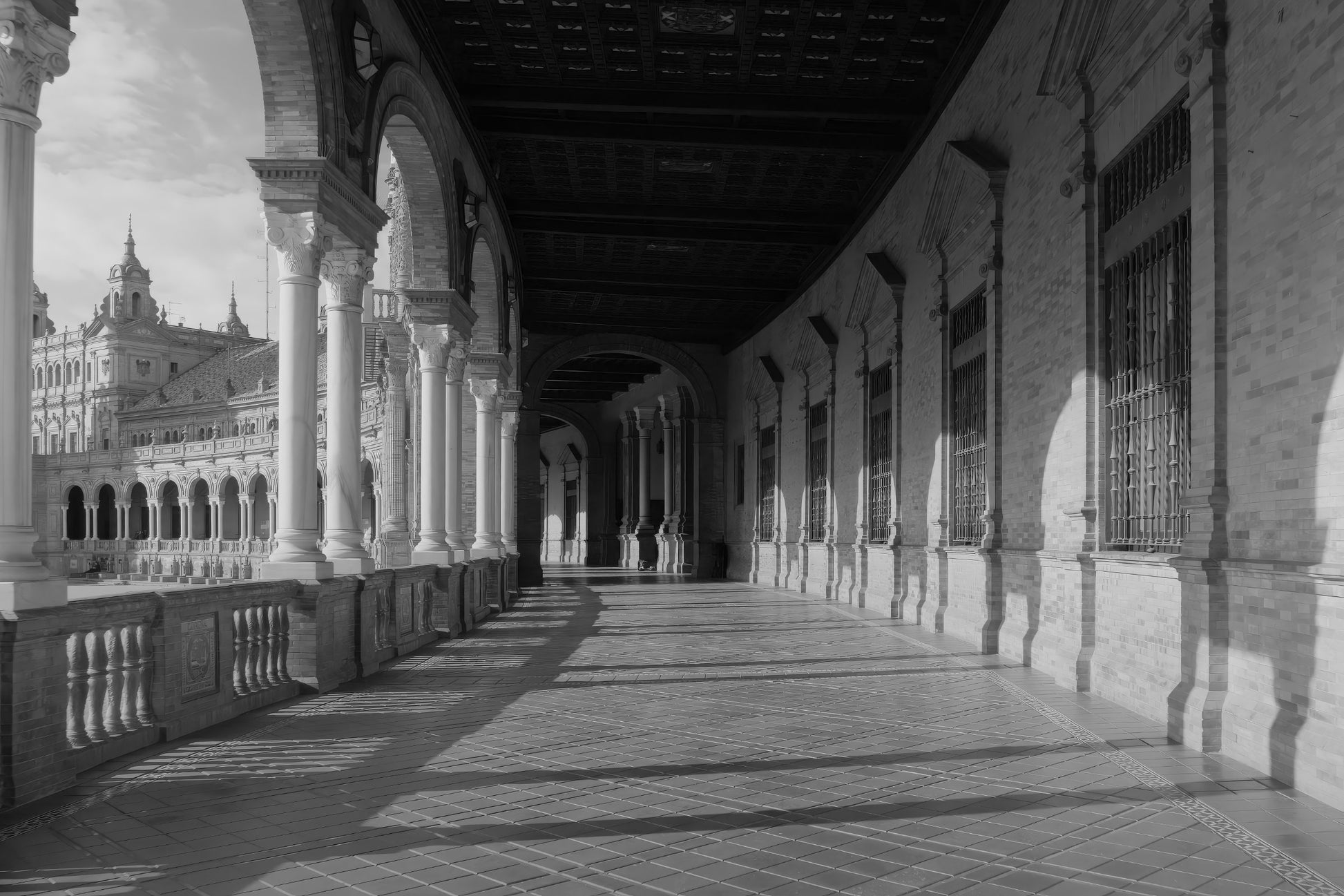 Black and white photograph of a grand architectural colonnade with arches, columns, tiled floor, and a distant ornate building.