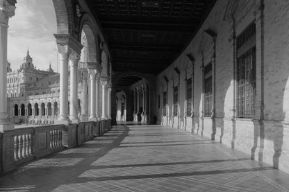 Black and white photograph of a grand architectural colonnade with arches, columns, tiled floor, and a distant ornate building.