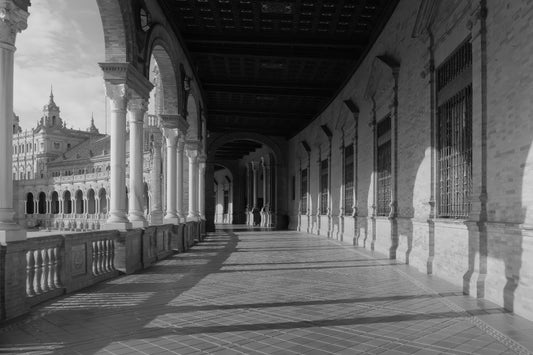 Black and white photograph of a grand architectural colonnade with arches, columns, tiled floor, and a distant ornate building.