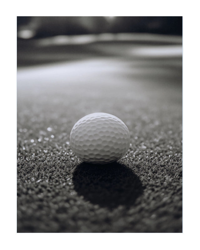 A single white golf ball resting on dark textured turf, photographed in a close-up outdoor-style shot with shallow depth of field.
