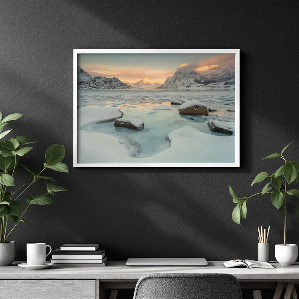 A framed art print depicting a snowy mountain landscape with a frozen lake at sunset, displayed on a dark gray wall above a styled desk with plants and books.