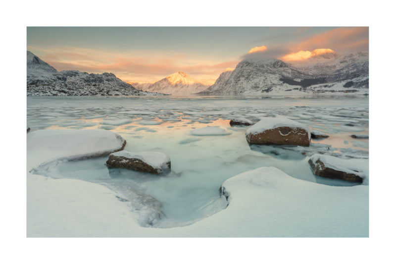 A scenic winter landscape print showing snow-covered rocks in a partially frozen icy body of water, with mountains and a soft sunset sky in the background.