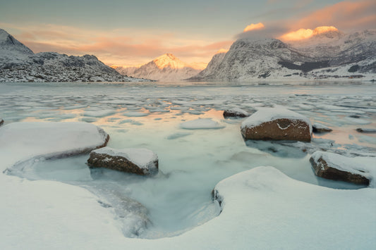 A snowy mountain landscape at sunset with a partially frozen lake in the foreground, rocks covered in snow, and warm light reflecting on the water and clouds.