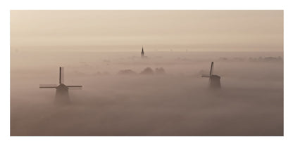 A misty landscape photograph featuring two dark windmills rising above thick fog, with a church spire faintly visible in the distance under a hazy beige sky.