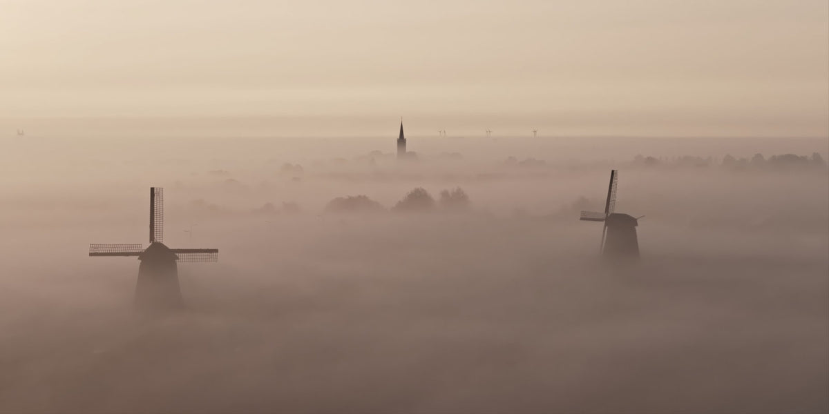 A misty landscape scene with two dark windmills rising above thick fog, and a church spire faintly visible in the distance under a pale hazy sky.