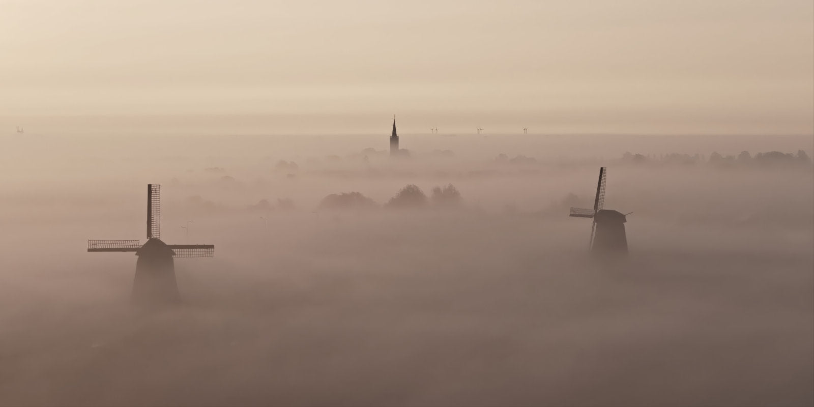 A misty landscape scene with two dark windmills rising above thick fog, and a church spire faintly visible in the distance under a pale hazy sky.