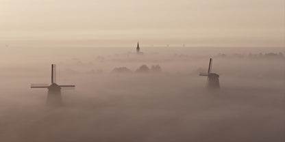 A misty landscape scene with two dark windmills rising above thick fog, and a church spire faintly visible in the distance under a pale hazy sky.