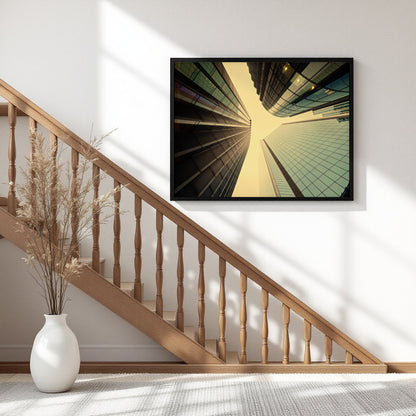 A framed wall art print depicting a dramatic low-angle view of modern skyscrapers converging toward the sky, displayed on a white wall above a wooden staircase in a bright, modern interior.