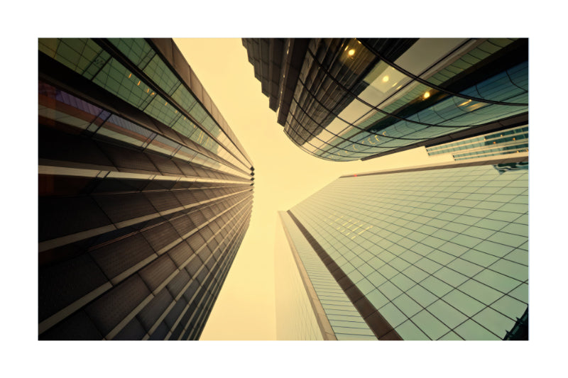 A low-angle photograph of modern skyscrapers with glass and dark metal facades converging toward the sky, presented with a white border.