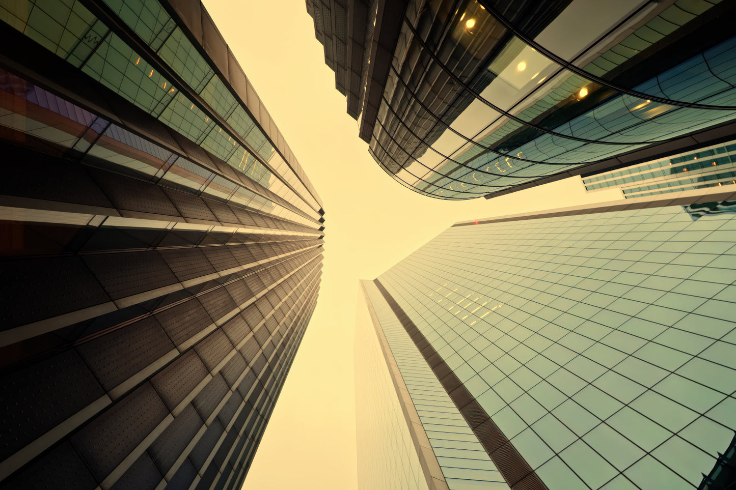 Low-angle horizontal upward view of modern glass skyscrapers framing a pale yellow sky between tall buildings.