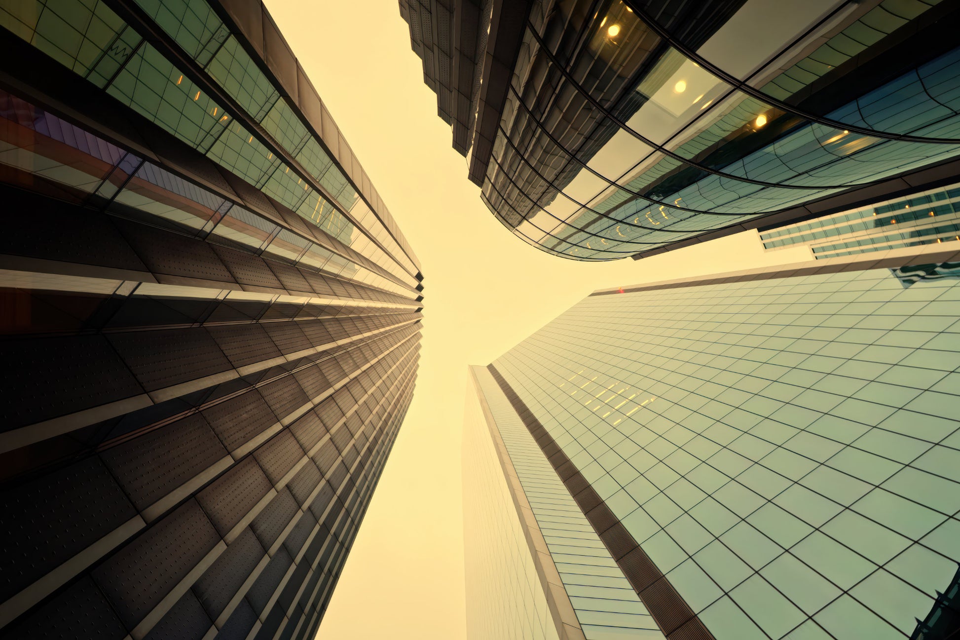 Low-angle horizontal upward view of modern glass skyscrapers framing a pale yellow sky between tall buildings.