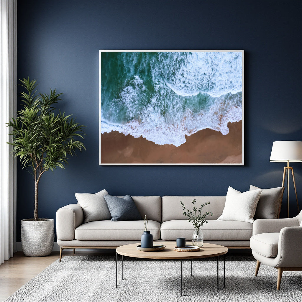 A large framed wall art print showing green and white ocean waves washing onto a brown beach, displayed above a light gray sofa in a modern living room with a dark blue wall.