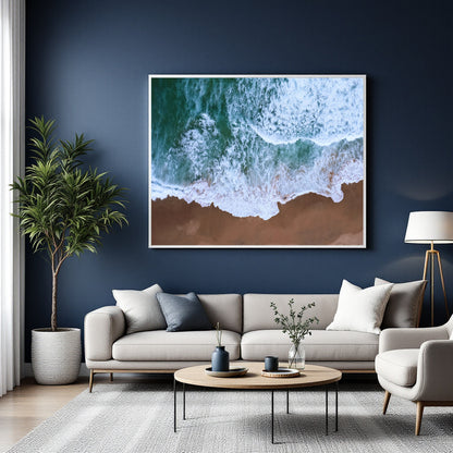 A large framed wall art print showing green and white ocean waves washing onto a brown beach, displayed above a light gray sofa in a modern living room with a dark blue wall.