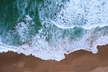 Aerial view of ocean waves with white foam crashing onto a sandy beach, showing turquoise-green water and tan shore.