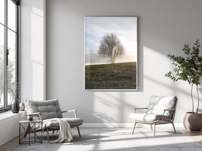 A vertical framed art print depicting a solitary tree on a grassy hill at sunrise, displayed on a light gray wall in a modern, sunlit living room with two gray armchairs and a potted plant.
