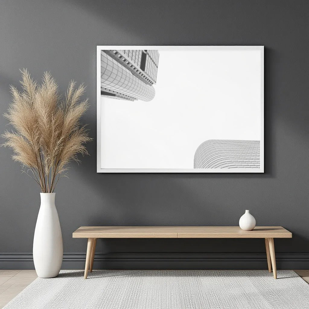 A minimalist framed wall art print featuring black and white architectural building lines on a white background, displayed on a dark gray wall above a light wood bench with a white vase of pampas grass and a small white vase.