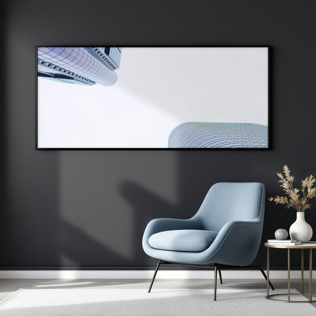 A large framed wall art print hangs on a dark charcoal wall above a light blue armchair, with a small side table and vase in the room. The artwork shows a minimalist architectural scene with blue curved building forms on a white background.