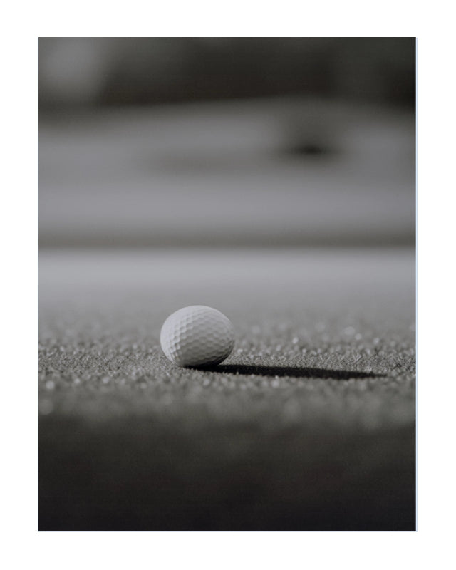 A white golf ball resting on a textured dark surface, photographed in a moody black-and-white style with a blurred background.