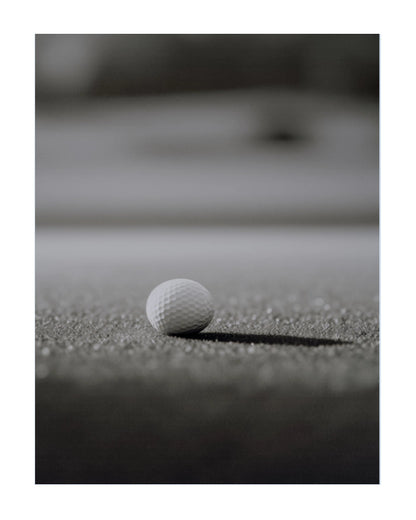 A white golf ball resting on a textured dark surface, photographed in a moody black-and-white style with a blurred background.