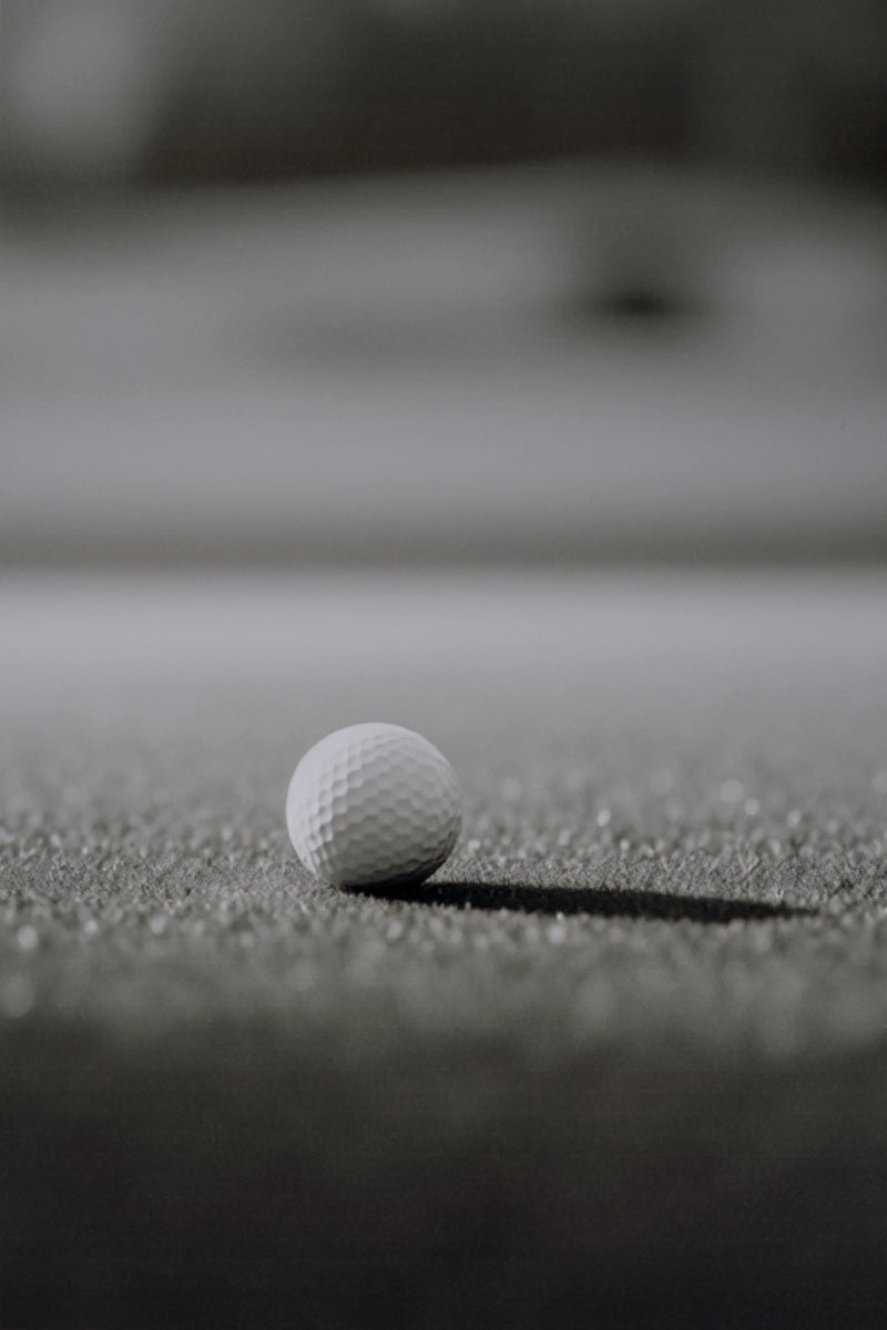 A single white golf ball resting on a dark textured surface, with a blurred gray background.