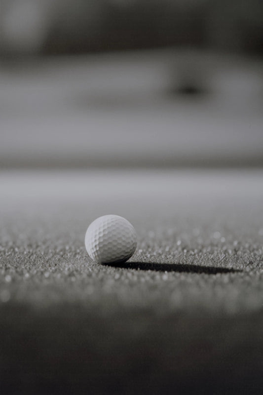 A single white golf ball resting on a dark textured surface, with a blurred gray background.