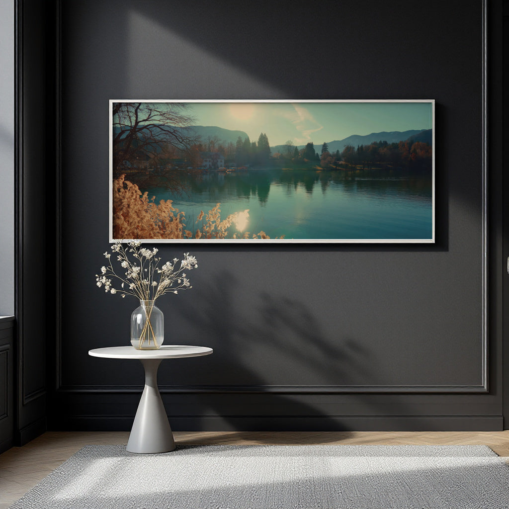 A panoramic framed landscape art print depicting a serene lake with mountains and autumn trees, displayed on a dark charcoal wall above a small white side table with a vase of white flowers.