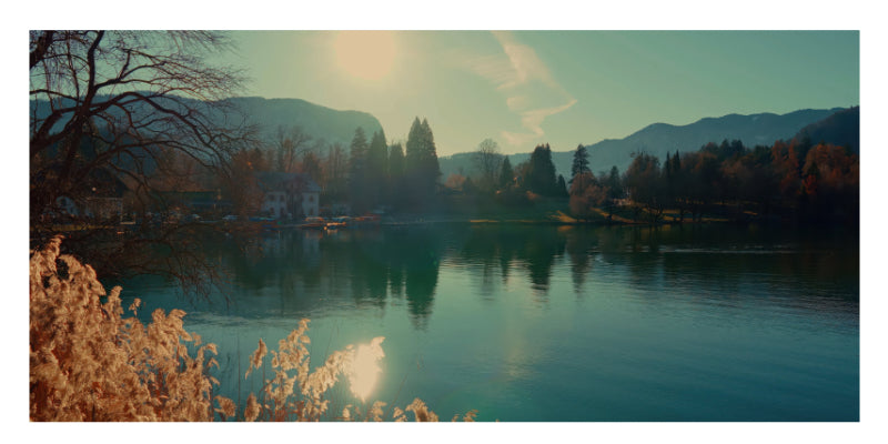A scenic landscape photograph of a calm lake with mountains in the background, trees along the shore, and warm sunlight reflecting on the water and foreground foliage.