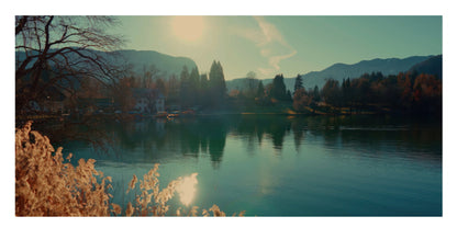 A scenic landscape photograph of a calm lake with mountains in the background, trees along the shore, and warm sunlight reflecting on the water and foreground foliage.