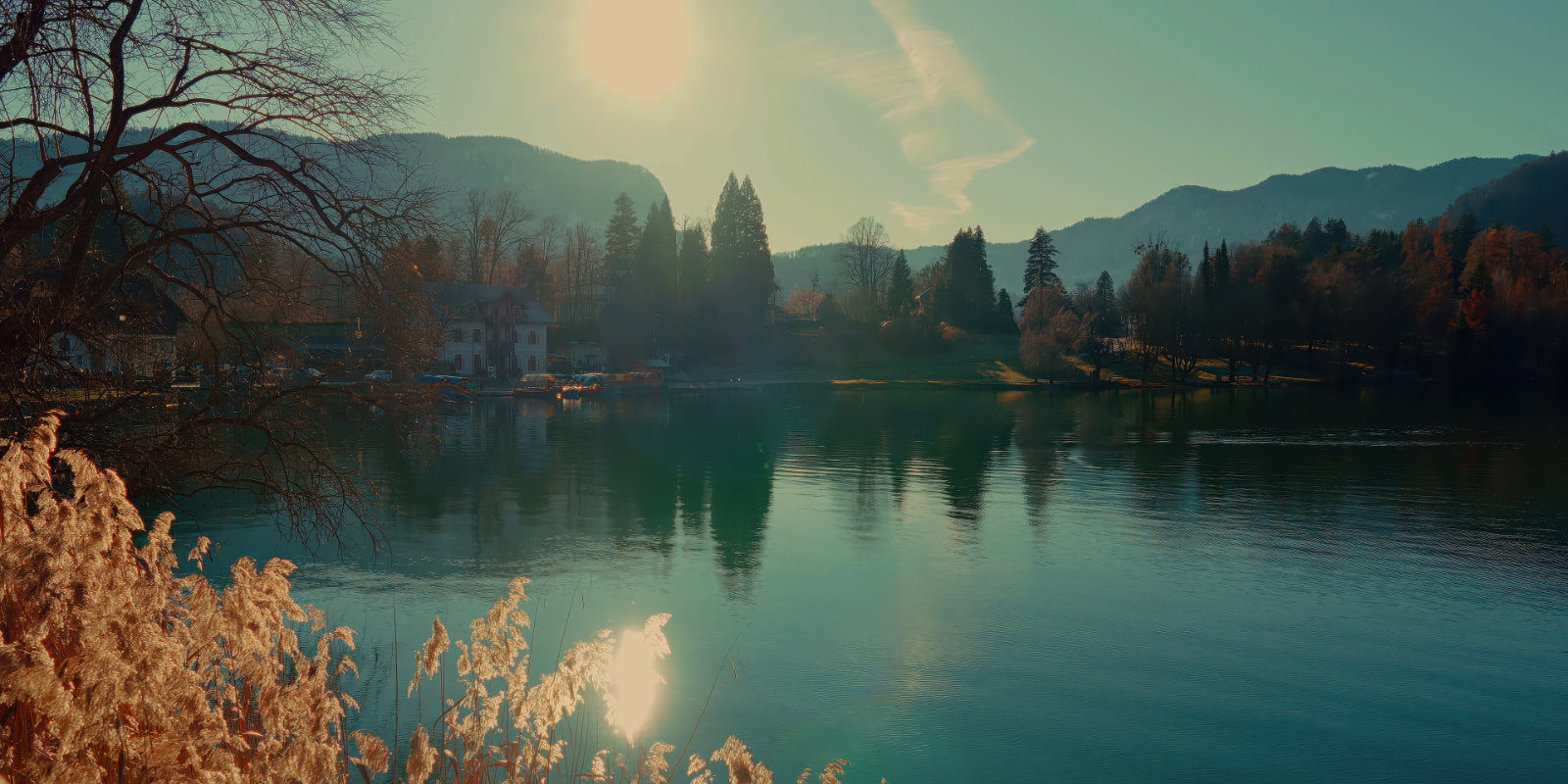 A scenic landscape photograph of a calm lake reflecting mountains, trees, and a bright sun, with autumn foliage in the foreground and distant shoreline buildings.