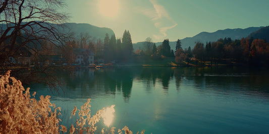 A scenic landscape photograph of a calm lake reflecting mountains, trees, and a bright sun, with autumn foliage in the foreground and distant shoreline buildings.