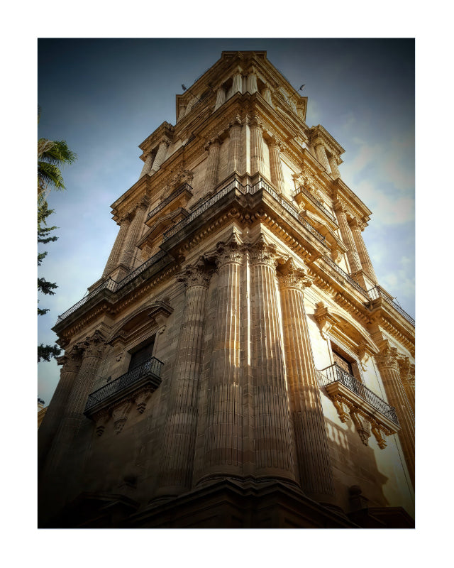 A low-angle photograph of a grand, historic stone tower with classical columns, set against a blue sky with a white border around the print.