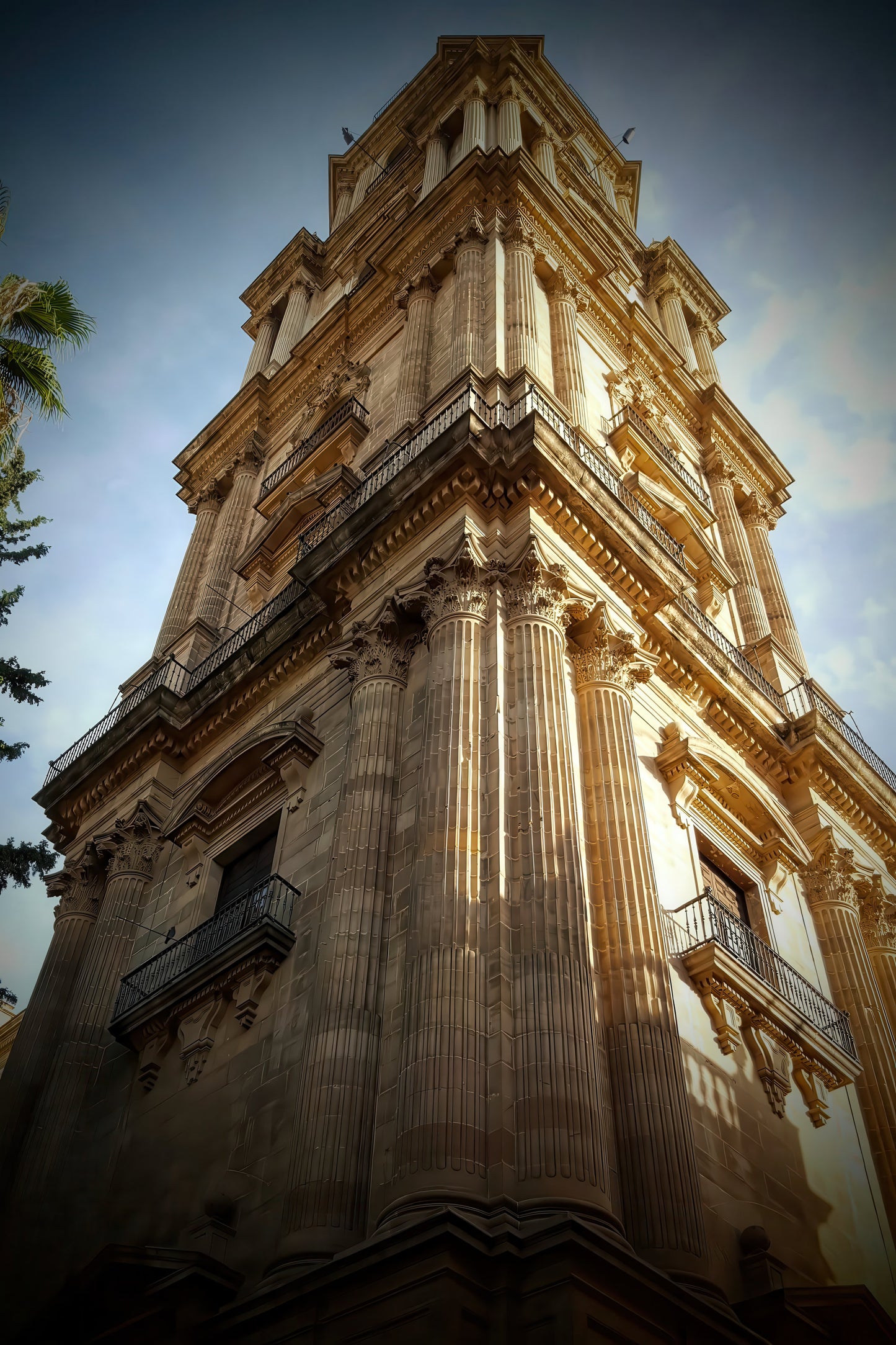 A low-angle photograph of a grand, historic stone tower with classical columns and balconies, set against a dramatic blue sky with warm sunlight highlighting its beige stone facade.
