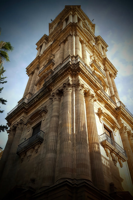 A low-angle photograph of a grand, historic stone tower with classical columns and balconies, set against a dramatic blue sky with warm sunlight highlighting its beige stone facade.
