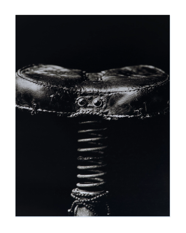 A black and white close-up photograph of a dark leather item with a stitched heart-shaped top and a coiled spring support, shown against a black background.