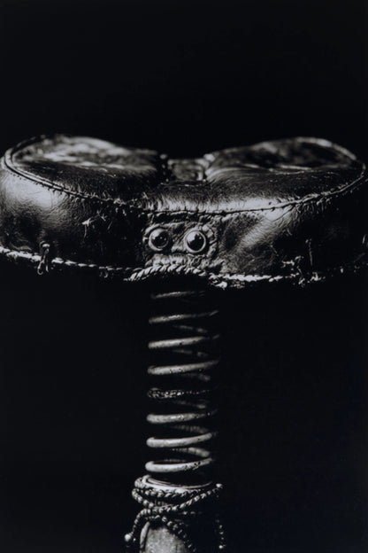 A close-up black and white photograph of a vintage leather saddle with a coiled spring underneath, set against a solid black background.