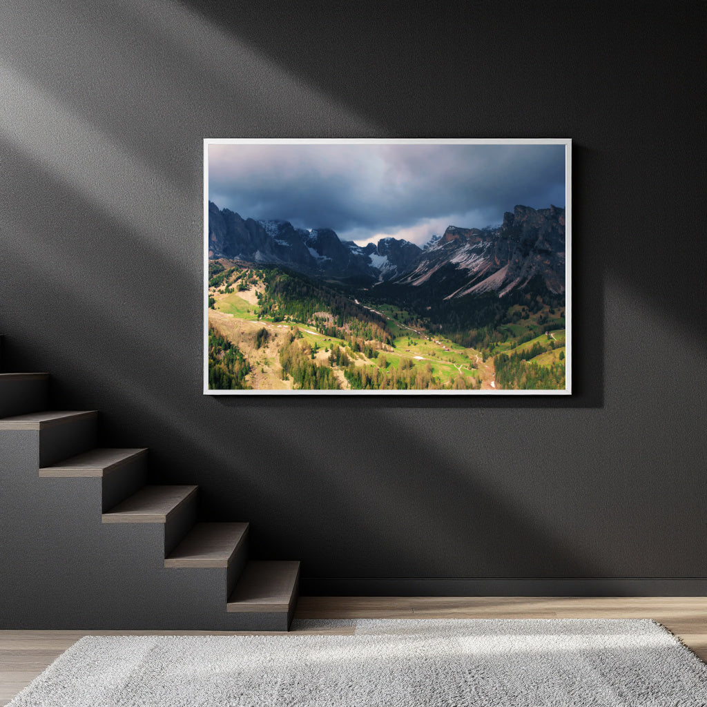 A framed landscape photograph of a dramatic mountain valley under stormy skies, displayed on a dark gray wall in a modern interior setting with a staircase and light beams.