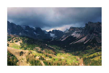 A scenic landscape photograph of a dramatic mountain range with dark storm clouds overhead, rocky peaks with patches of snow, and a green valley below with trees and winding paths.