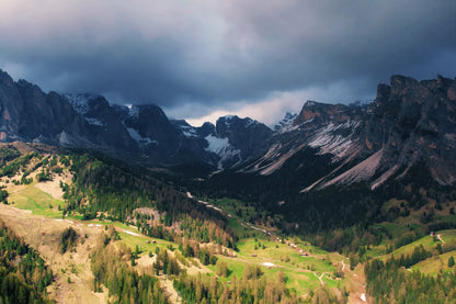 A dramatic mountain landscape with steep rocky peaks, dark storm clouds overhead, patches of snow, and green valley meadows with trees and winding paths.