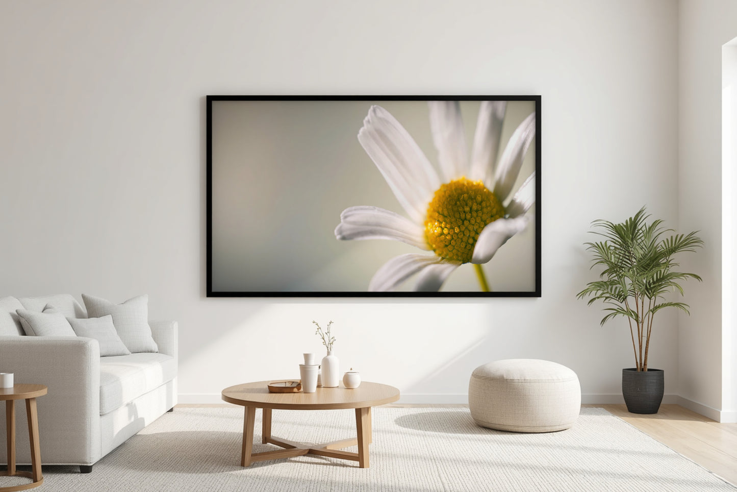 A large framed wall art print depicting a close-up white daisy with a yellow center, displayed in a modern living room setting above a light wood coffee table and next to a potted plant.