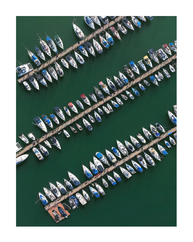 Aerial view of multiple sailboats moored along diagonal docks over deep green water.
