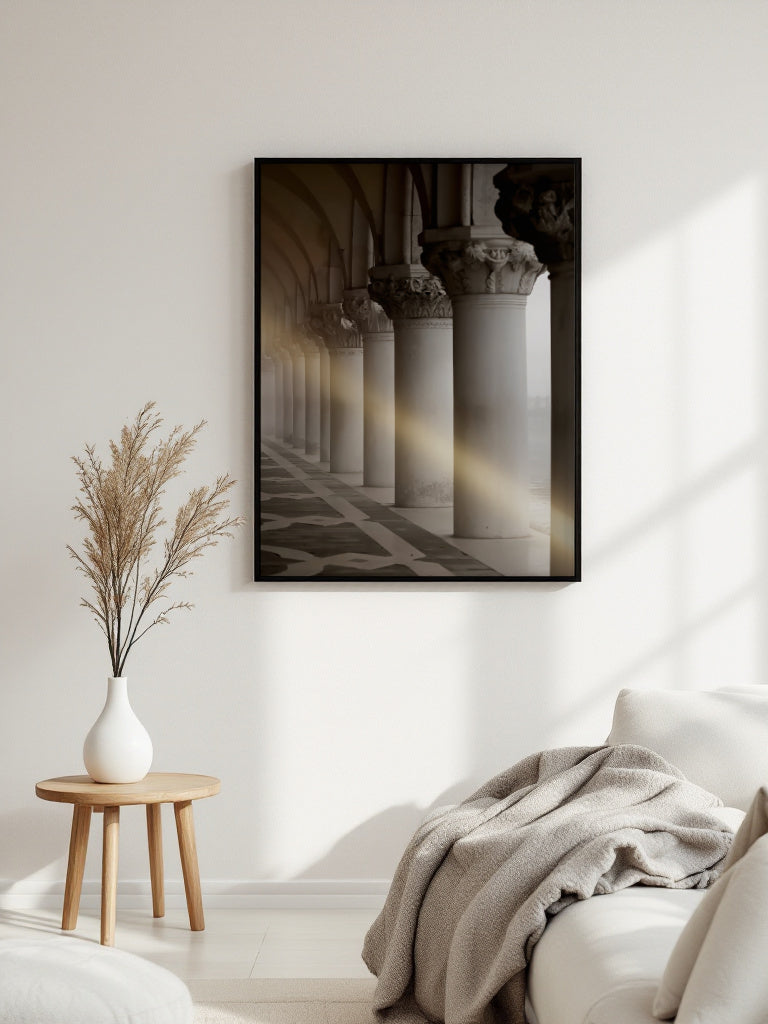 A framed black-and-white architectural art print depicting classical columns and arches with warm sunlight streaming through, displayed on a white wall in a modern living room setting next to a wooden side table with a vase of dried grass and a sofa with a throw blanket.