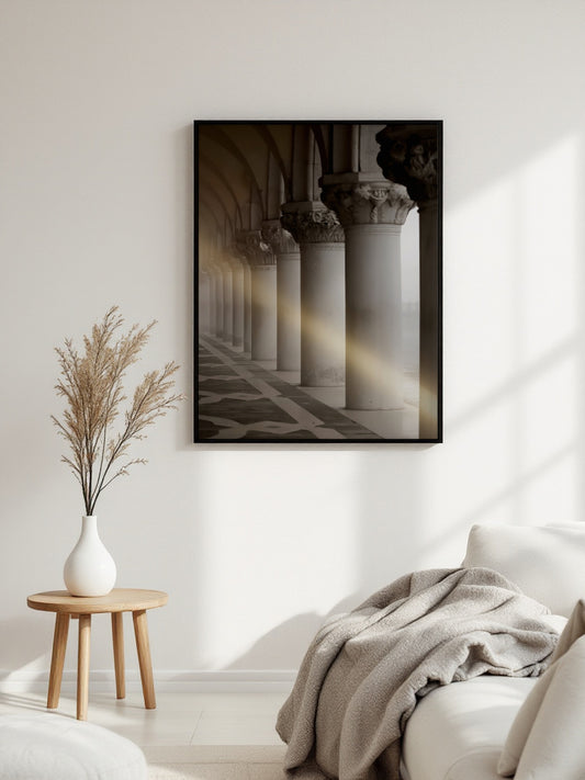 A framed black-and-white architectural art print depicting classical columns and arches with warm sunlight streaming through, displayed on a white wall in a modern living room setting next to a wooden side table with a vase of dried grass and a sofa with a throw blanket.