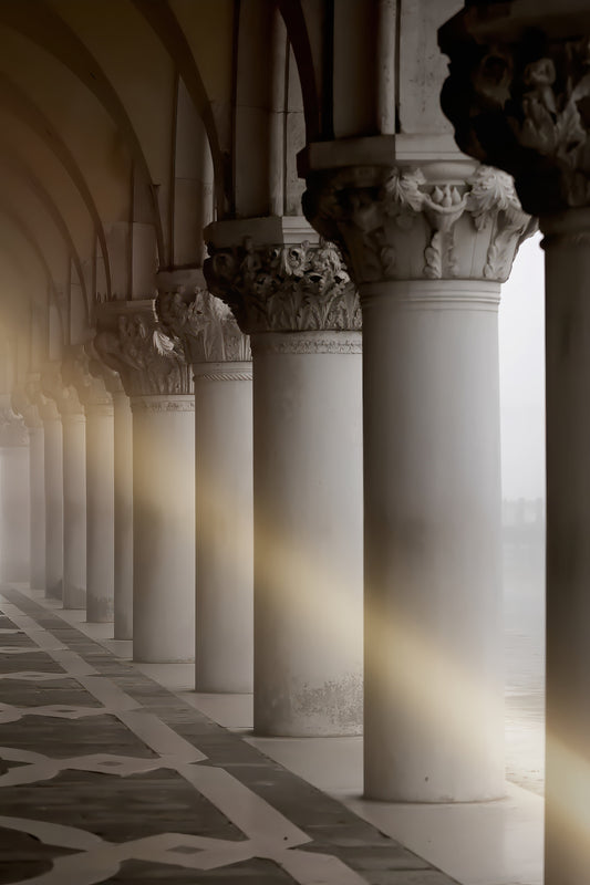 A scenic photograph of a grand colonnade with tall white columns, detailed capitals, and warm sunlight streaming through the arches onto a patterned stone floor.