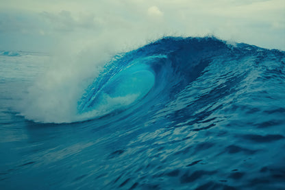 A dramatic blue ocean wave curls into a barrel with white foam at the crest under an overcast sky.