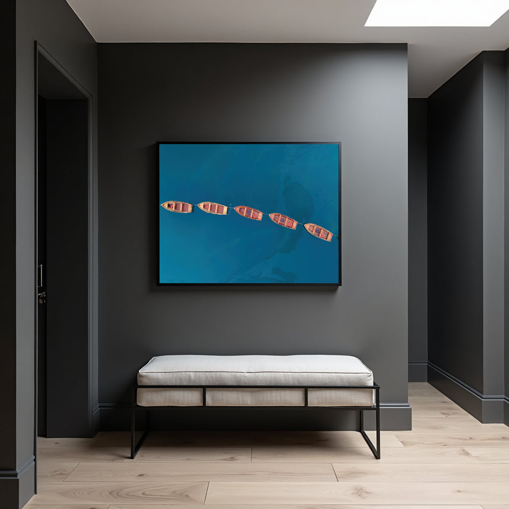 A framed wall art print showing four red-and-white striped boats on a bright blue water background, displayed above a light upholstered bench in a modern dark gray entryway with wood flooring.