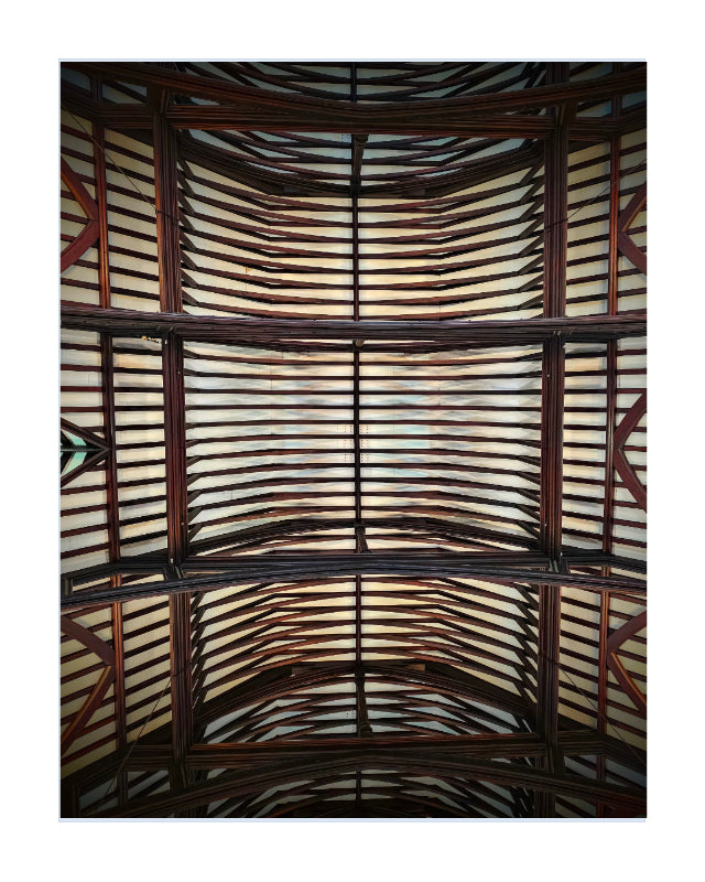 An upward-looking photograph of a large arched wooden ceiling or pavilion roof, featuring dark brown timber beams and numerous light-colored, warm-toned horizontal panels or slats.