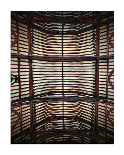 An upward-looking photograph of a large arched wooden ceiling or pavilion roof, featuring dark brown timber beams and numerous light-colored, warm-toned horizontal panels or slats.